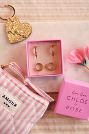 Coffret de parure de bijoux Marie contenant deux paires de mini créoles dorées striées - Bijoux photographiés dans un écrin à bijoux rose avec des accessoires et décorations roses et dorées