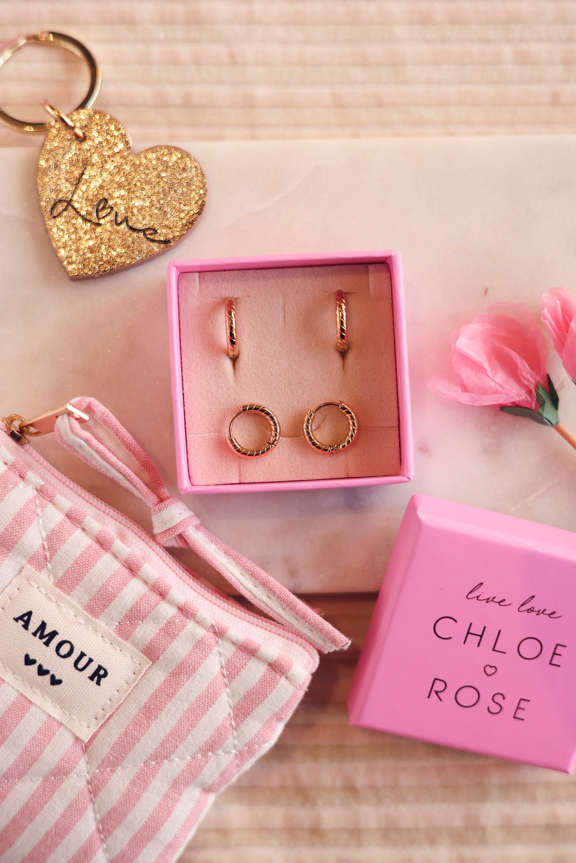 Coffret de parure de bijoux Marie contenant deux paires de mini créoles dorées striées - Bijoux photographiés dans un écrin à bijoux rose avec des accessoires et décorations roses et dorées