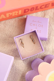 Coffret de parure de bijoux Divine contenant deux paires de mini créoles dorées ornées de zircons - Bijoux photographiés dans un écrin à bijoux lila posé dans une ambiance colorée