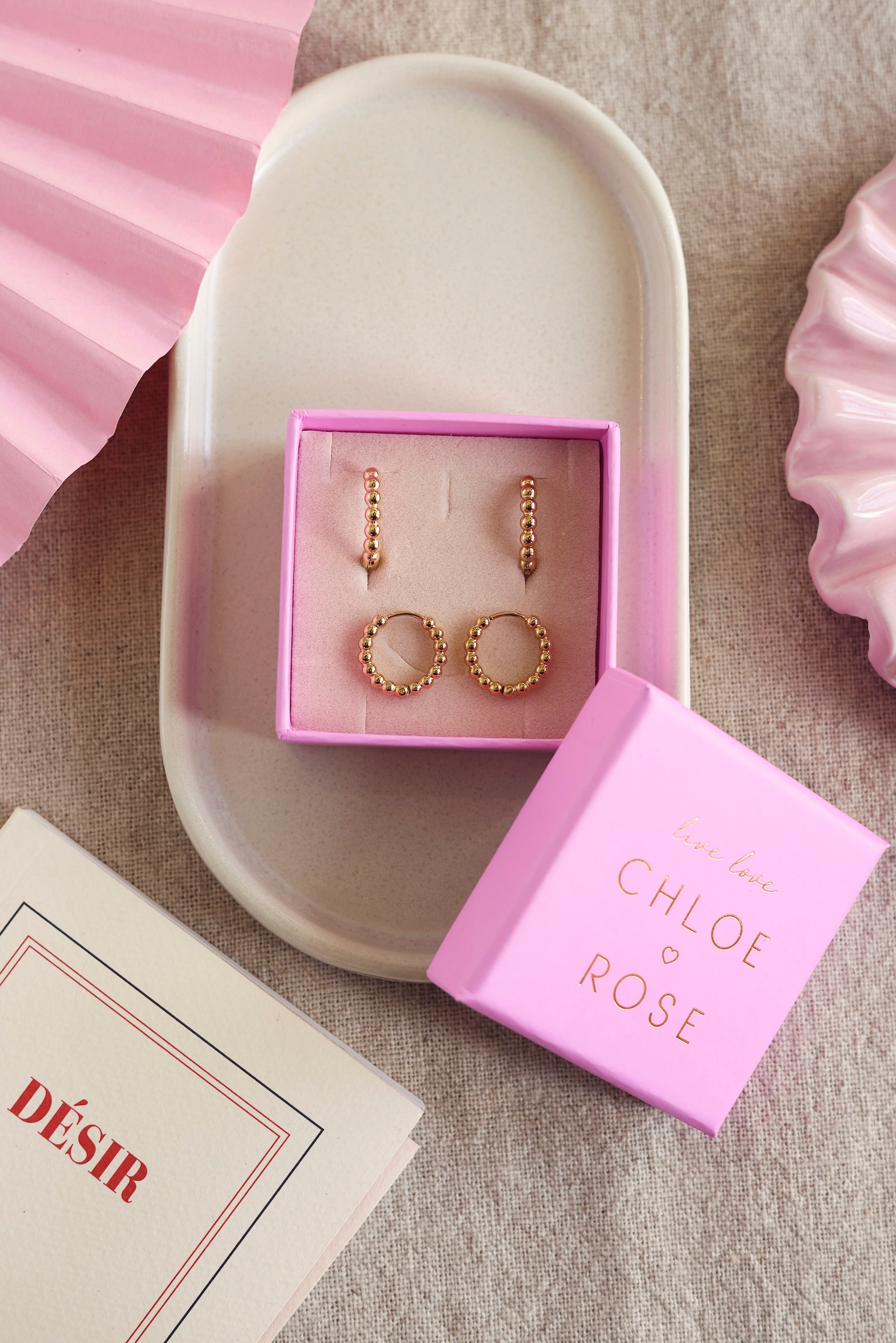 Coffret de parure de bijoux Laperla contenant deux paires de mini créoles dorées perlées - Bijoux photographiés dans un écrin à bijoux rose dans une coupole blanche avec des décorations roses