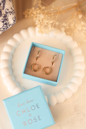Coffret de parure de bijoux Laura contenant deux paires de mini créoles dorées torsadées - Bijoux photographiés dans un écrin à bijoux bleu posé sur un vide poche blanc avec des fleurs séchées