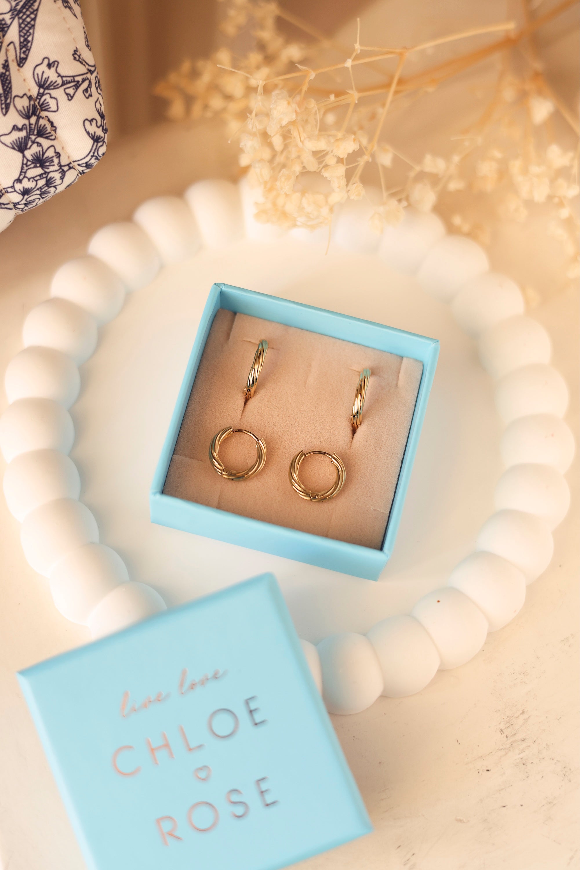 Coffret de parure de bijoux Laura contenant deux paires de mini créoles dorées torsadées - Bijoux photographiés dans un écrin à bijoux bleu posé sur un vide poche blanc avec des fleurs séchées