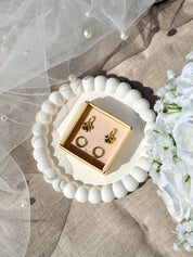 Coffret de parure de bijoux Lola comprenant une paire de mini créoles dorées ornées d’une fleur ainsi qu’une paire de créoles simples - Coffret photographié dans une coupole blanche avec un voile de mariée et un bouquet