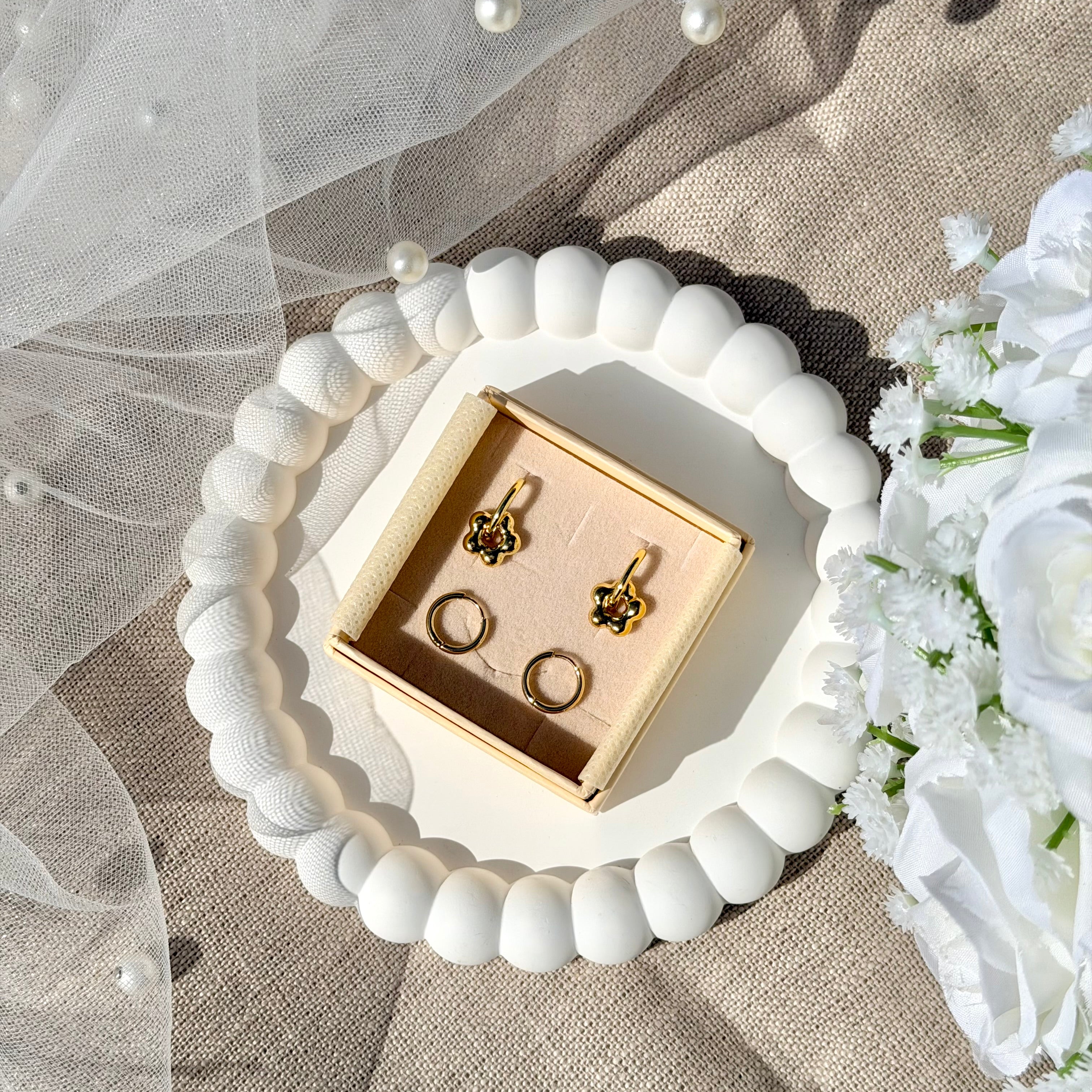 Coffret de parure de bijoux Lola comprenant une paire de mini créoles dorées ornées d’une fleur ainsi qu’une paire de créoles simples - Coffret photographié dans une coupole blanche avec un voile de mariée et un bouquet