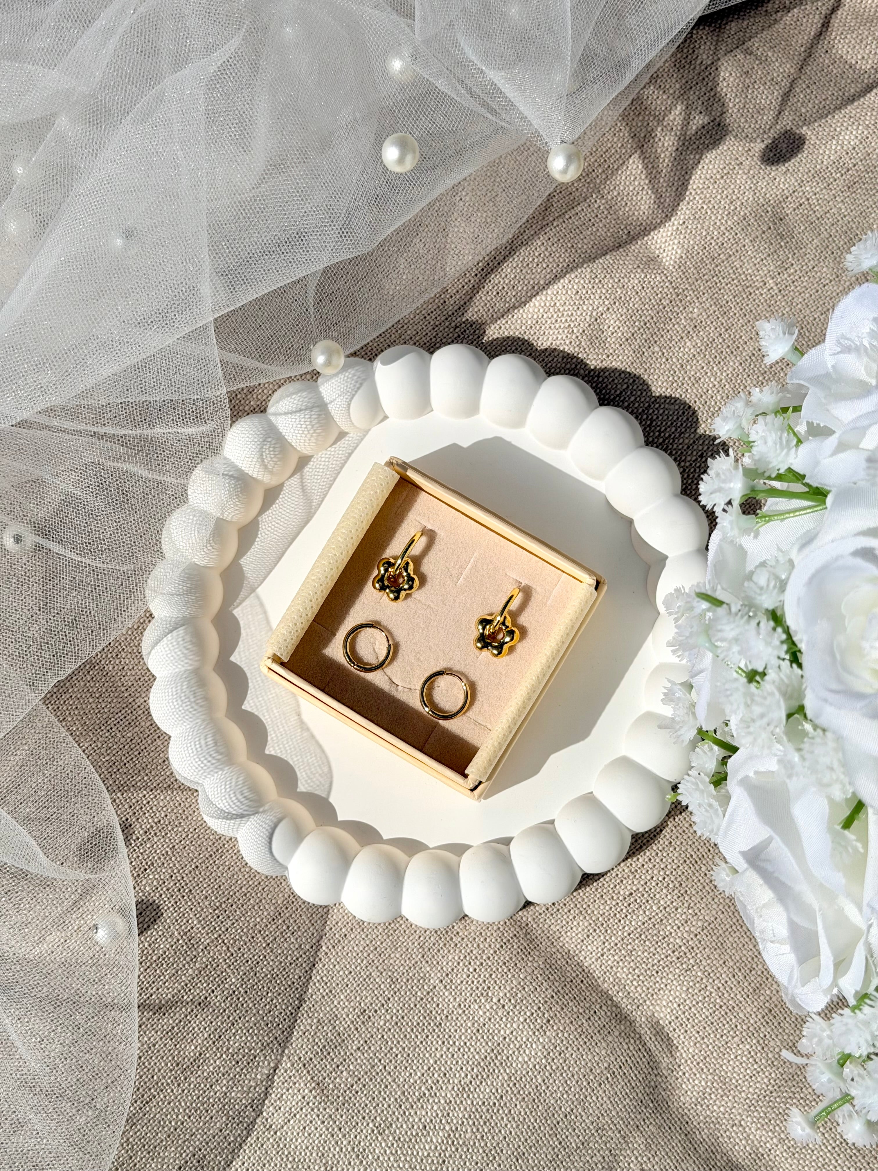 Coffret de parure de bijoux Lola comprenant une paire de mini créoles dorées ornées d’une fleur ainsi qu’une paire de créoles simples - Coffret photographié dans une coupole blanche avec un voile de mariée et un bouquet