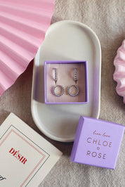 Coffret de parure de bijoux Laperla contenant deux paires de mini créoles dorées perlées - Bijoux photographiés dans un écrin à bijoux violet dans une coupole blanche avec des décorations roses