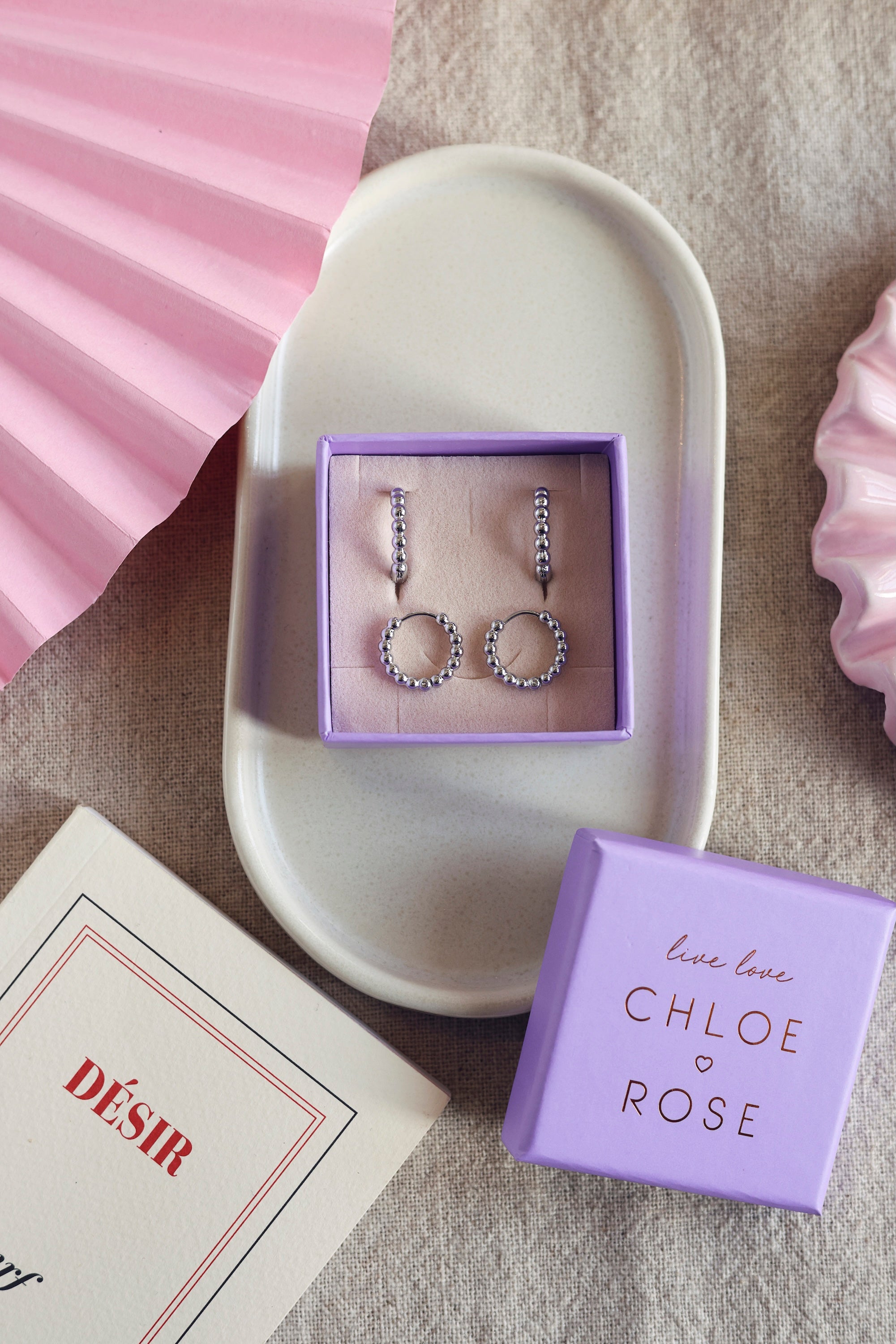 Coffret de parure de bijoux Laperla contenant deux paires de mini créoles dorées perlées - Bijoux photographiés dans un écrin à bijoux violet dans une coupole blanche avec des décorations roses
