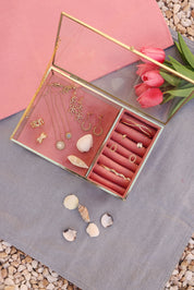 Collier doré Buffy aigue marine soleil - Bijou photographié dans une boite à bijou en velours rose et en verre aux bords doré posé sur un tissu bleu posé sur des cailloux avec des tulipes et des coquillages