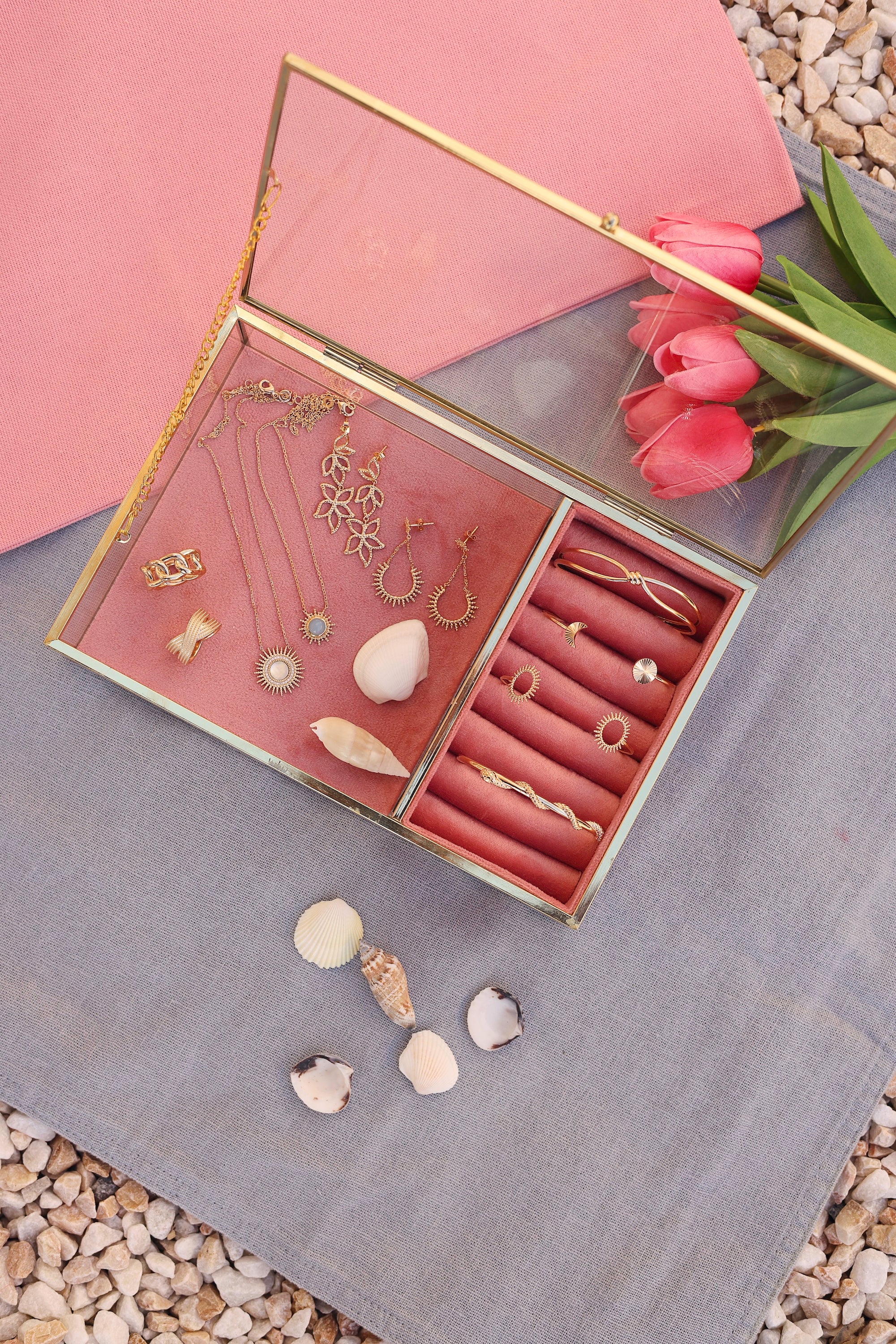 Collier doré Buffy aigue marine soleil - Bijou photographié dans une boite à bijou en velours rose et en verre aux bords doré posé sur un tissu bleu posé sur des cailloux avec des tulipes et des coquillages