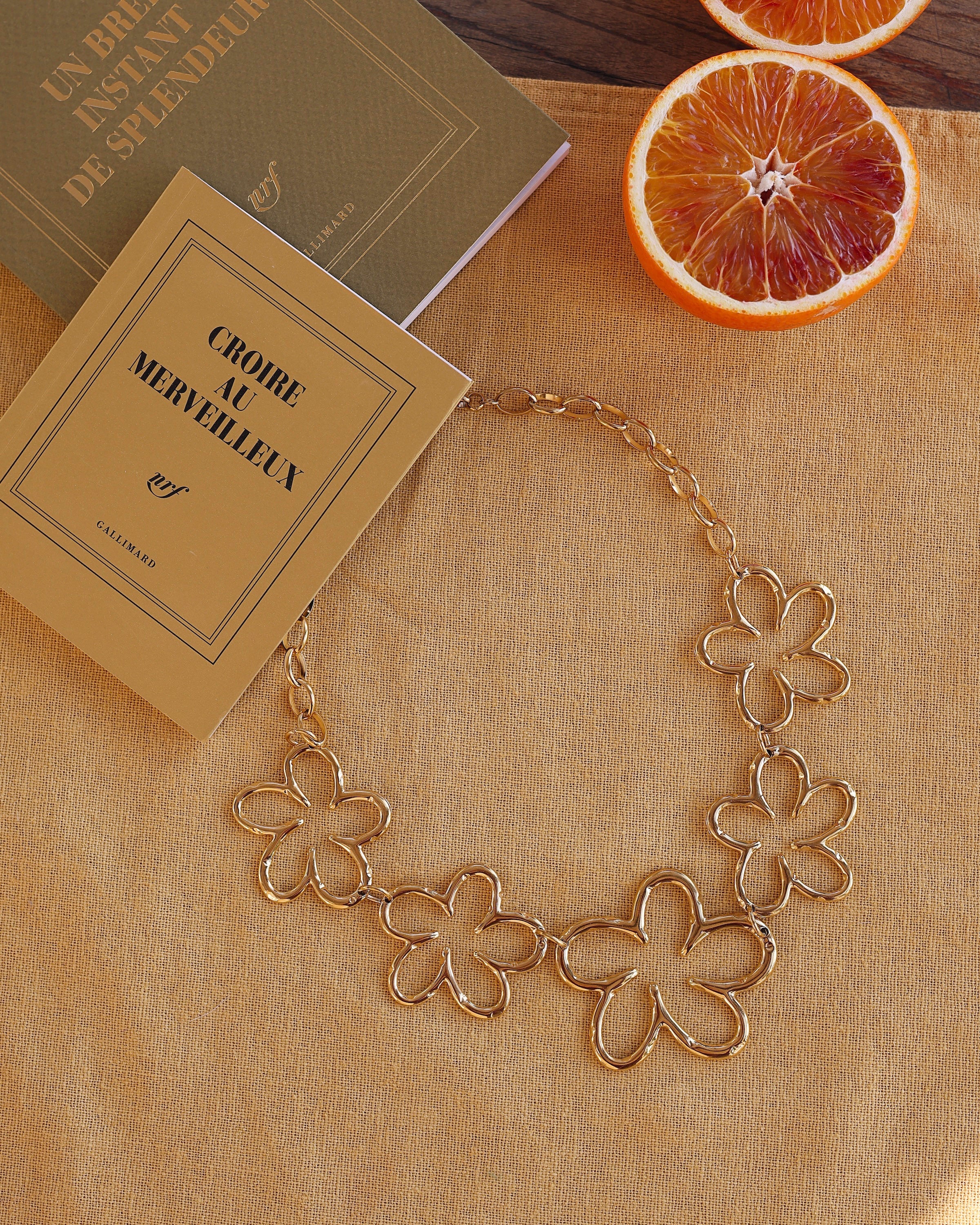 Collier statement doré Oksana fleurs - Bijou photographié sur tissu jaune avec livres et oranges sanguines