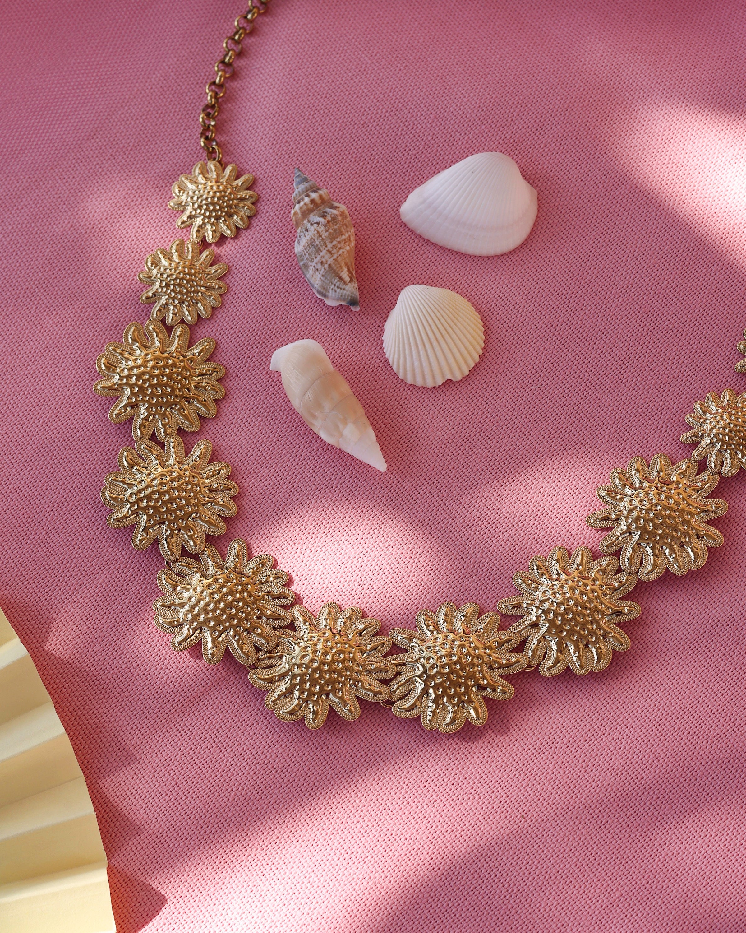 Collier doré Darina avec motifs floraux en relief - Bijou photographié sur un tissu rose avec des coquillages