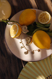 Collier doré Talyssa avec pendentifs tortue, soleil et coquillages - bijou photographié sur assiette blanche avec citrons et boucles d'oreilles coquillages
