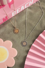 Collier personnalisable Rita en argent avec médaille au contour orné de motifs délicatement ajourés et sertis de zircons - Bijou photographié sur un tissu kaki avec des décorations roses