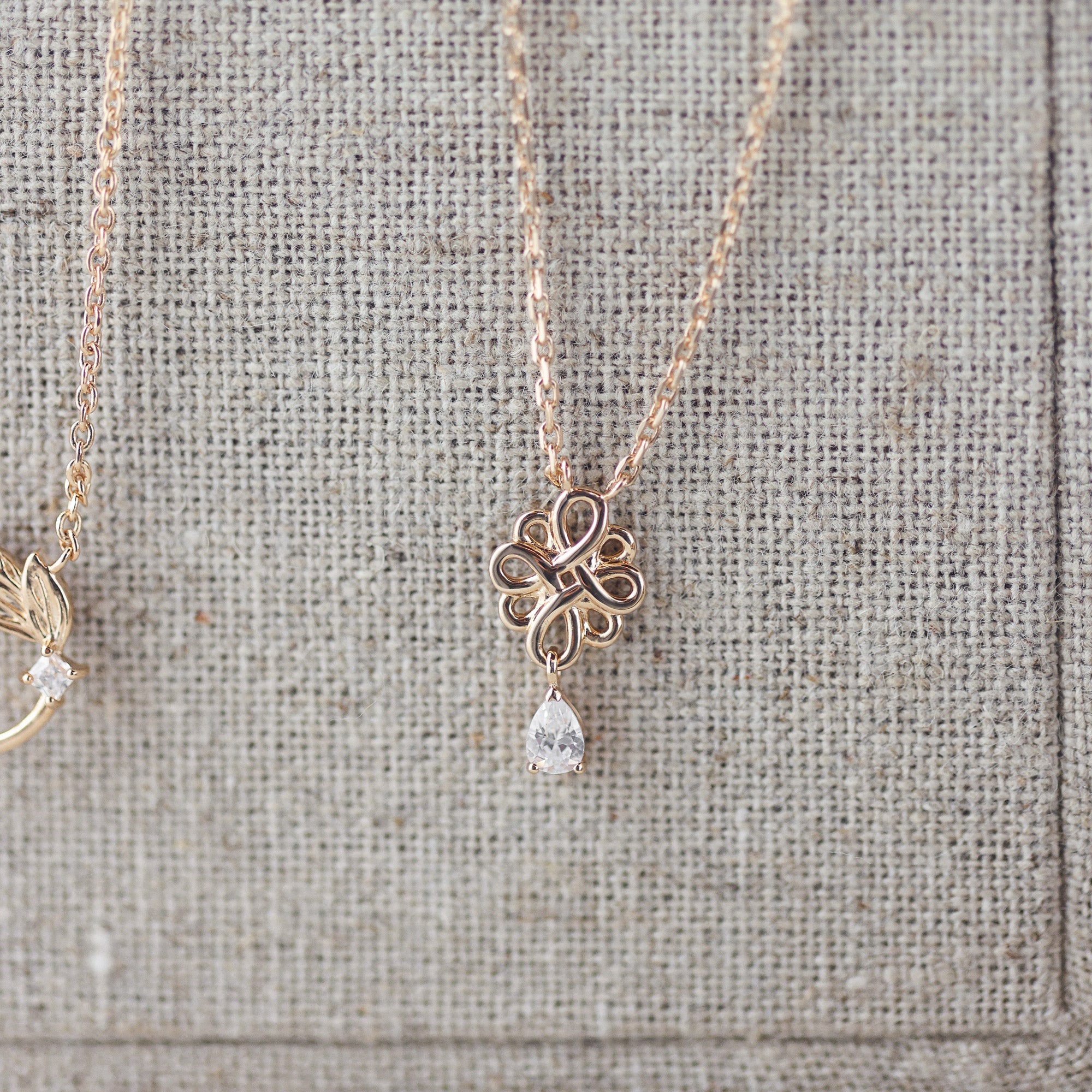 Collier doré nœud de l’infini avec pendentif zircon Joanie - Bijou photographié sur présentoir en lin gris