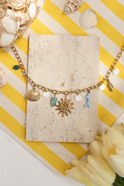 Collier maille doré Maja avec pendentifs coquillages, corail, soleil et pierres naturelles - Bijou photographié dans un décor estival