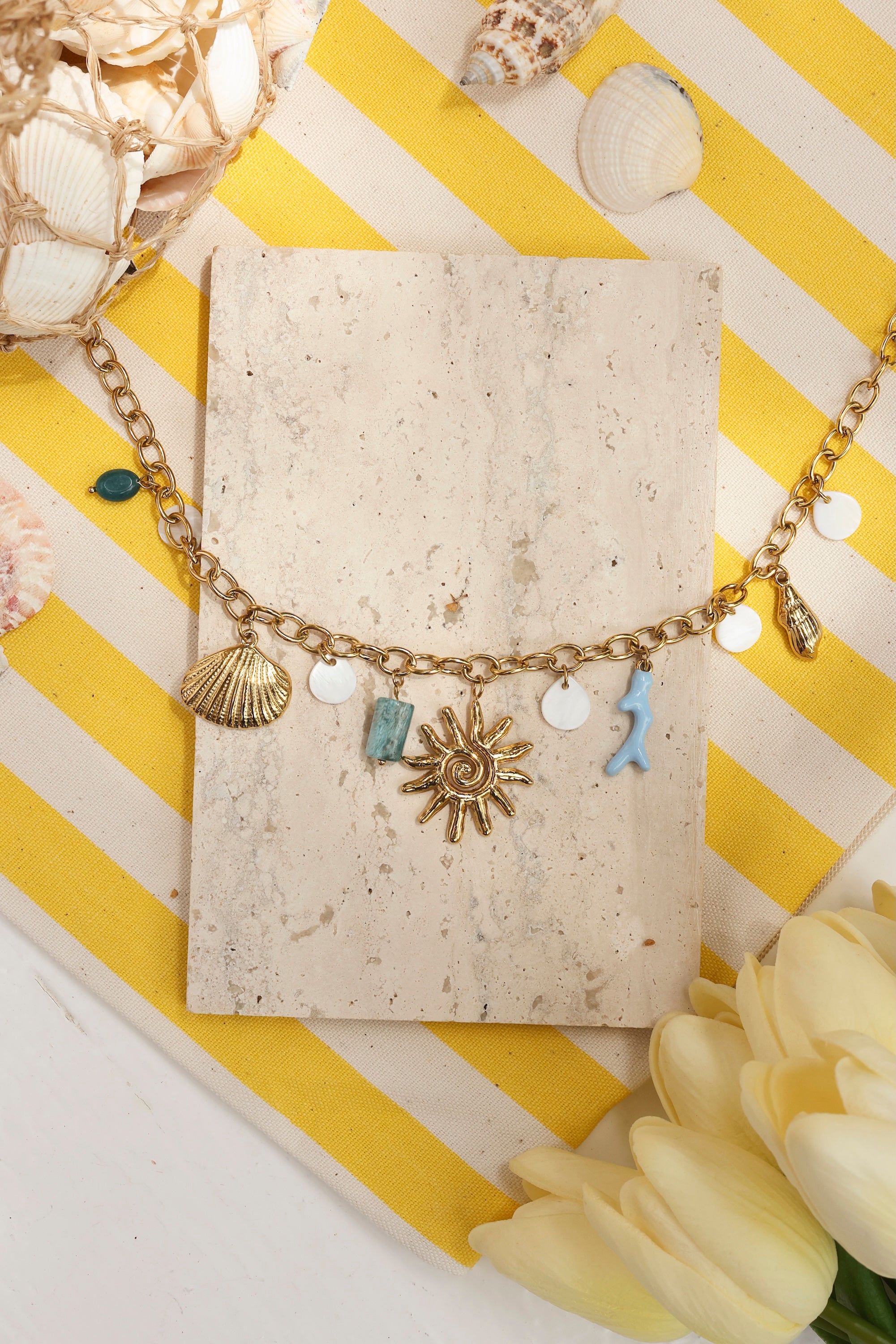 Collier maille doré Maja avec pendentifs coquillages, corail, soleil et pierres naturelles - Bijou photographié dans un décor estival