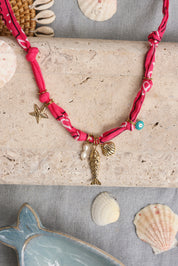 Collier doré Yarina rose tissu bandana rose avec pendentifs étoile de mer, nacres, poisson, coquillage et œil - Bijou photographié dans un décor estival
