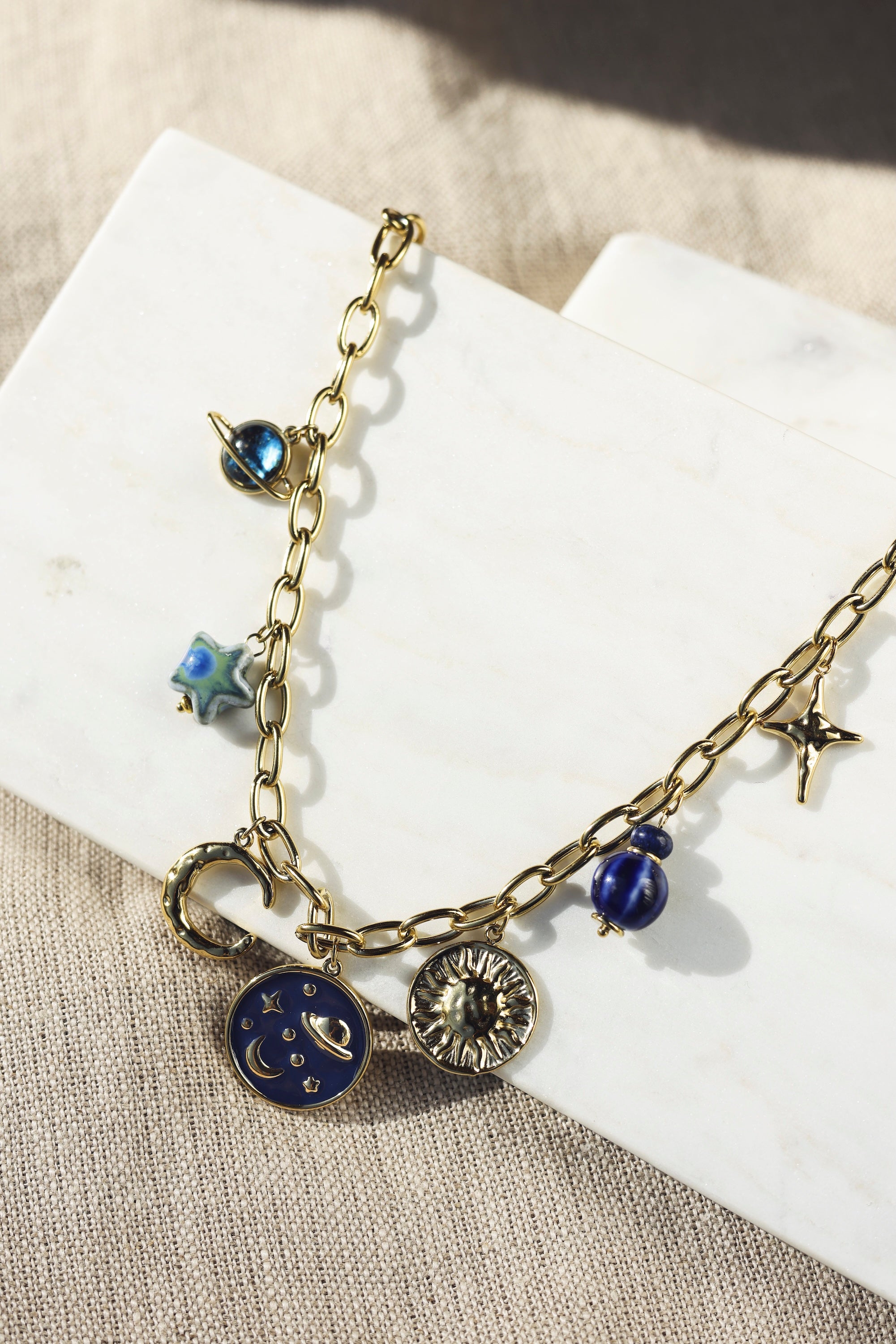 Collier en maille doré Alyssa avec pendentifs lunes, soleil, planètes et étoiles - Bijou photographié sur un présentoir en marbre blanc posé sur un tissu en lin beige