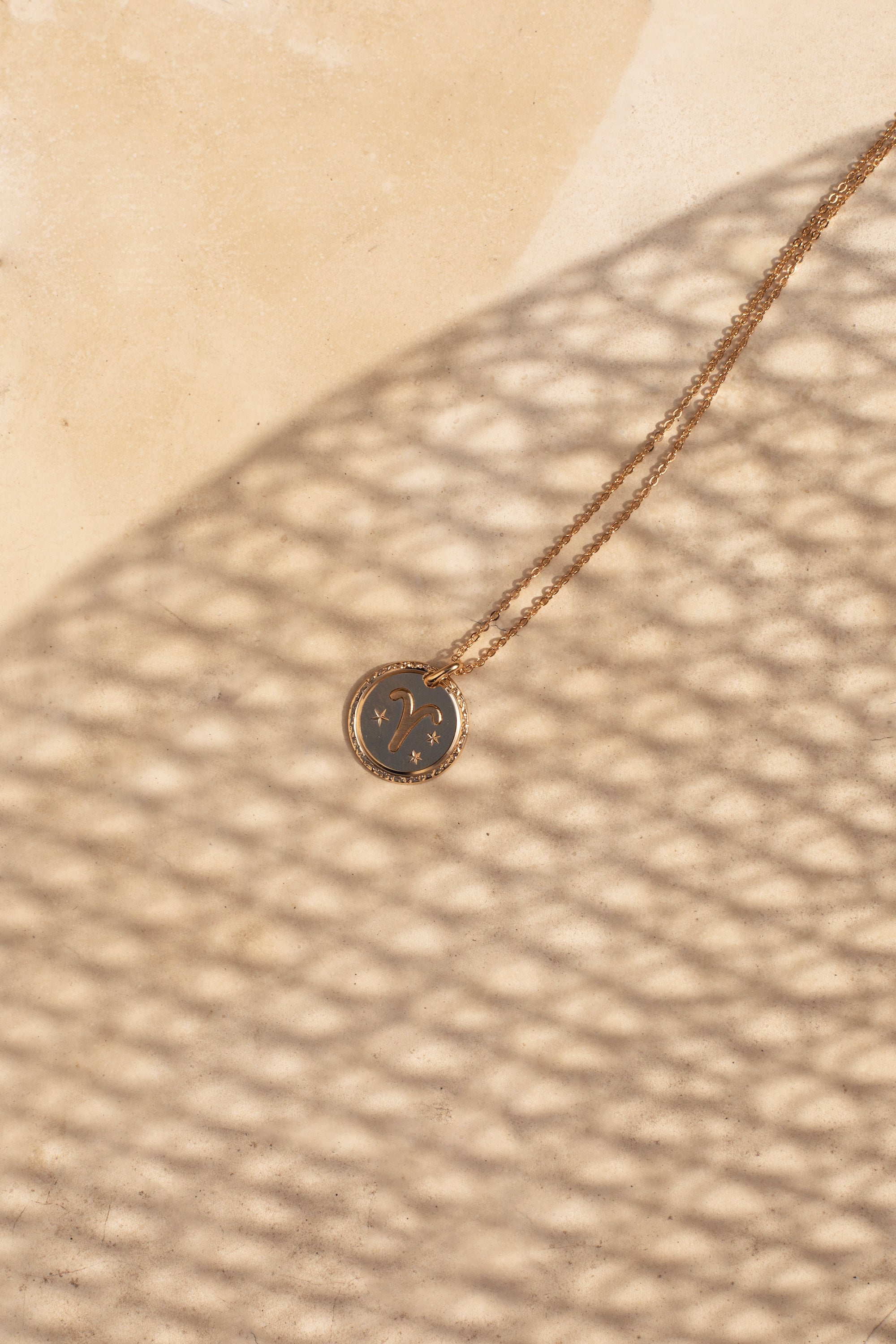  Collier doré signe du zodiaque Bélier - Bijou photographié dans un jeu d'ombre sur de la pierre beige