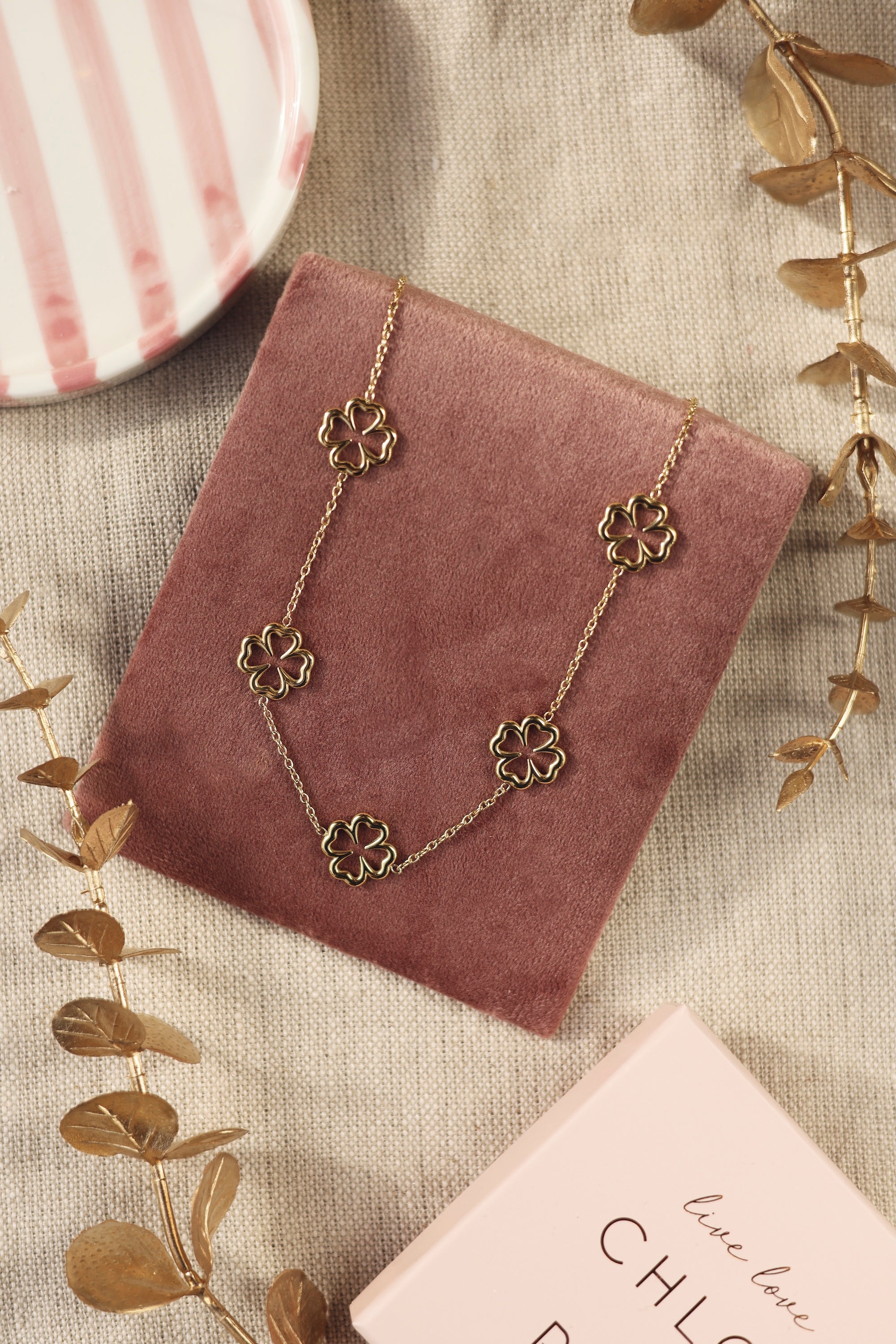 Collier doré Colline trèfles - Bijou photographié sur un présentoir en velours rose foncé posé sur un tissu en lin avec des feuilles décoratives dorées