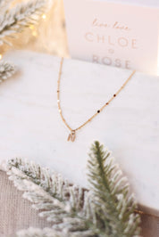 Collier personnalisable doré Laora chaîne ornée de petites pastilles pour lettres de l'alphabet et charms - Bijou photographié sur du marbre blanc avec des branches de sapin