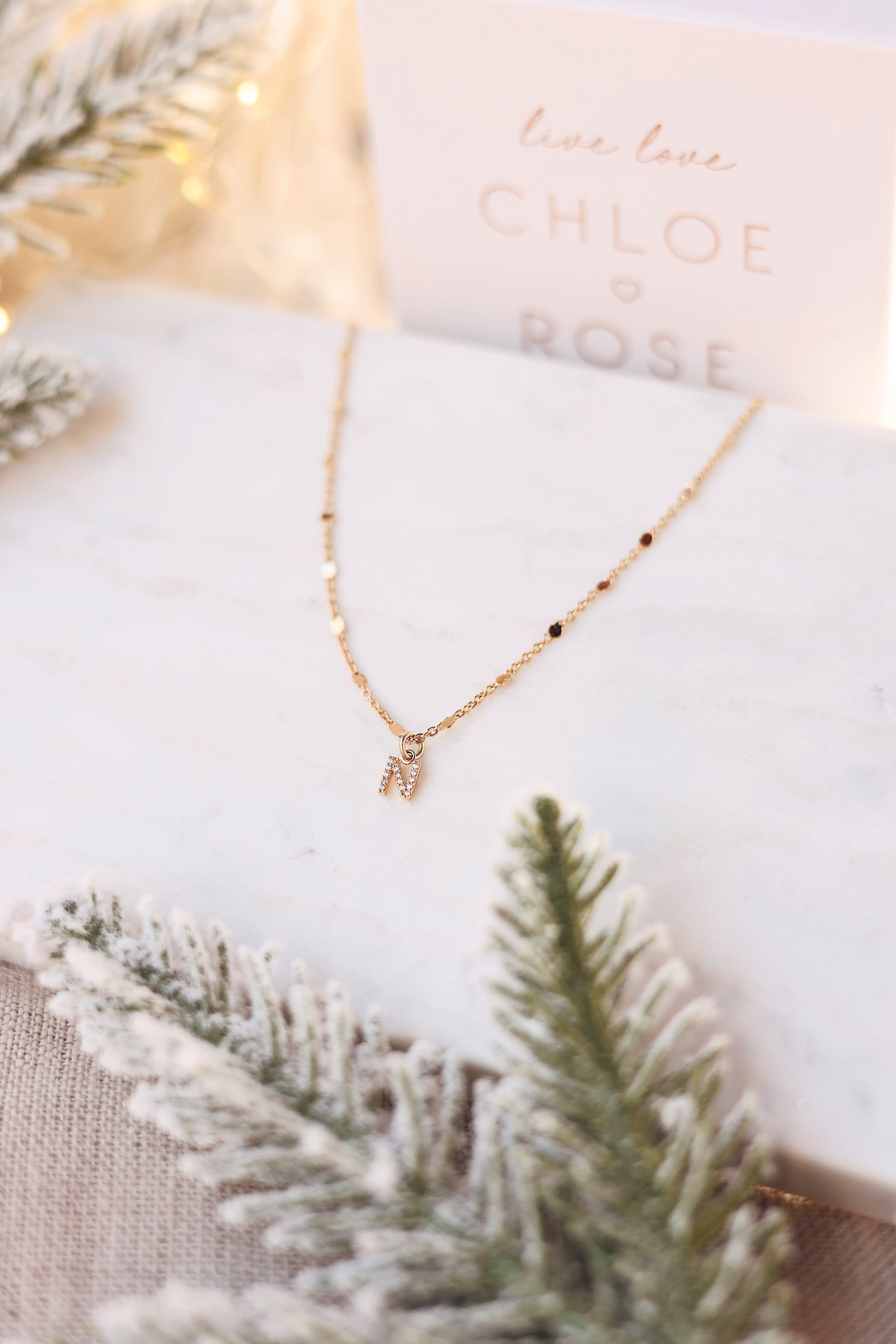 Collier personnalisable doré Laora chaîne ornée de petites pastilles pour lettres de l'alphabet et charms - Bijou photographié sur du marbre blanc avec des branches de sapin
