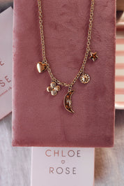 Collier doré Lorna en maille avec pendentifs lune, fleur, soleil, coeur et étoile - Bijou photographié sur un présentoir en velours rose foncé