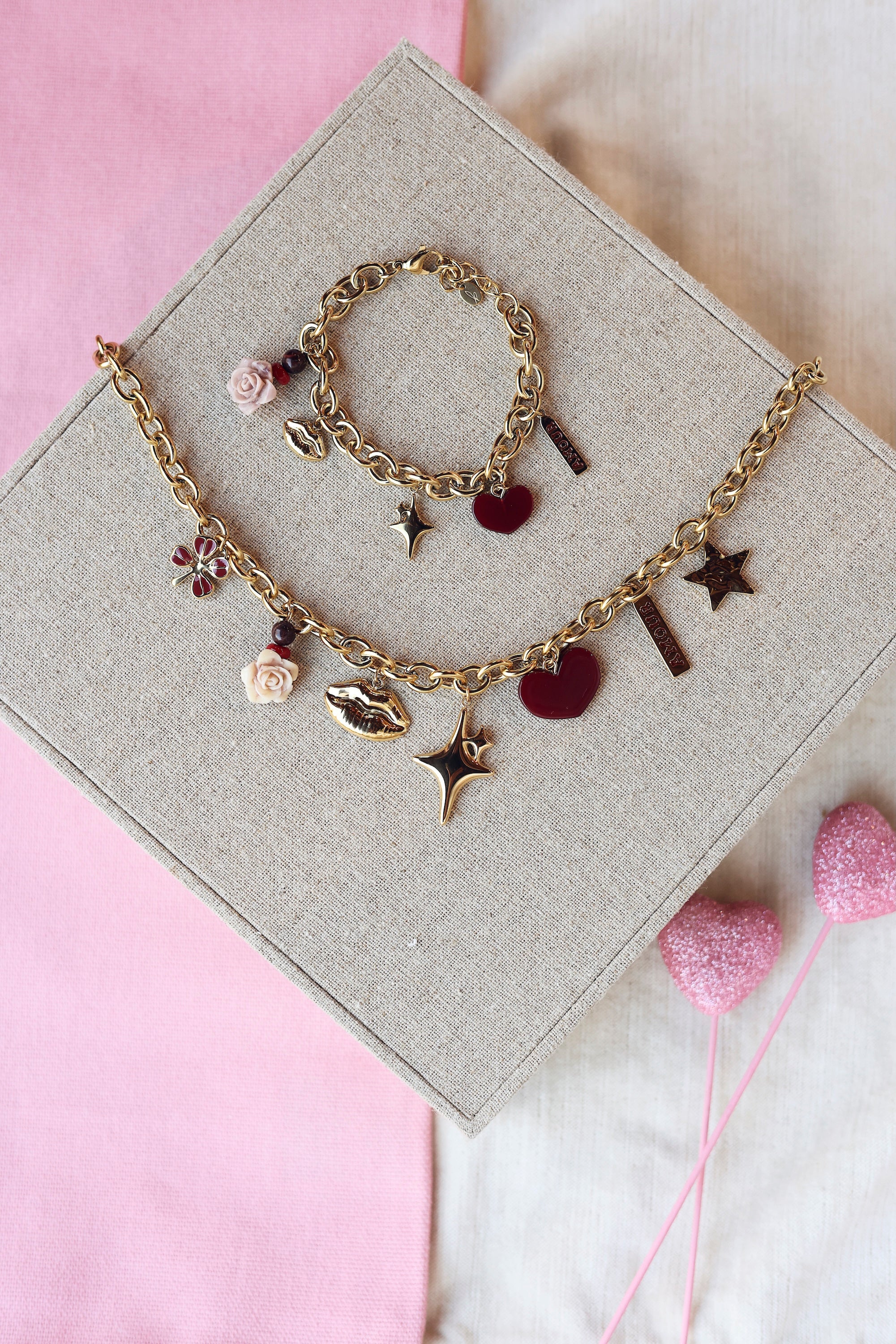 Collier doré Swanne maille avec pendentifs trèfle, rose, bouche, étoile, coeur et&nbsp;amulette avec inscription "amour" - Bijou photographié sur un présentoir en lin gris
