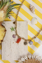 Collier maille doré Melania avec pendentifs coquillages, poissons, étoile de mer, soleil et coeur - Bijou photographié dans un décor estival