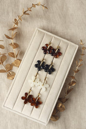 Créoles dorées Shauna marron avec pendentif fleur en acétate - Bijou photographié sur un présentoir blanc crème posé sur un tissu en lin avec des feuilles décoratives dorées