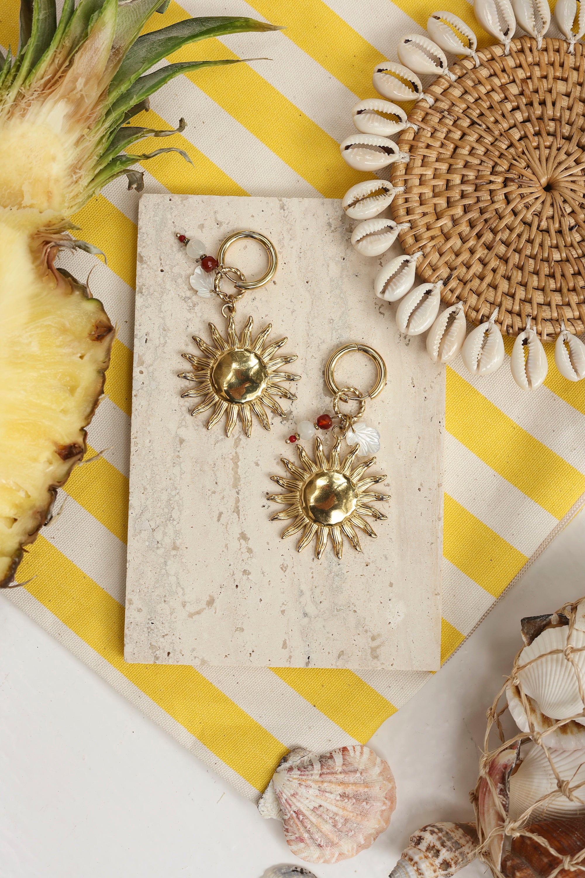 Boucles d'oreilles mini créoles dorées Natanya avec pendentifs soleil, feuille en nacre et pierres naturelles - Bijou photographié dans un décor estival