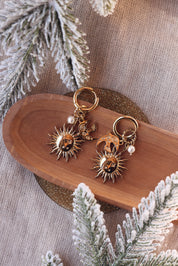 Boucles d'oreilles mini créoles dorées Solenia pendentif soleil, lune, nacre et hirondelle - Bijou photographié sur une coupole en bois avec des branches de sapin