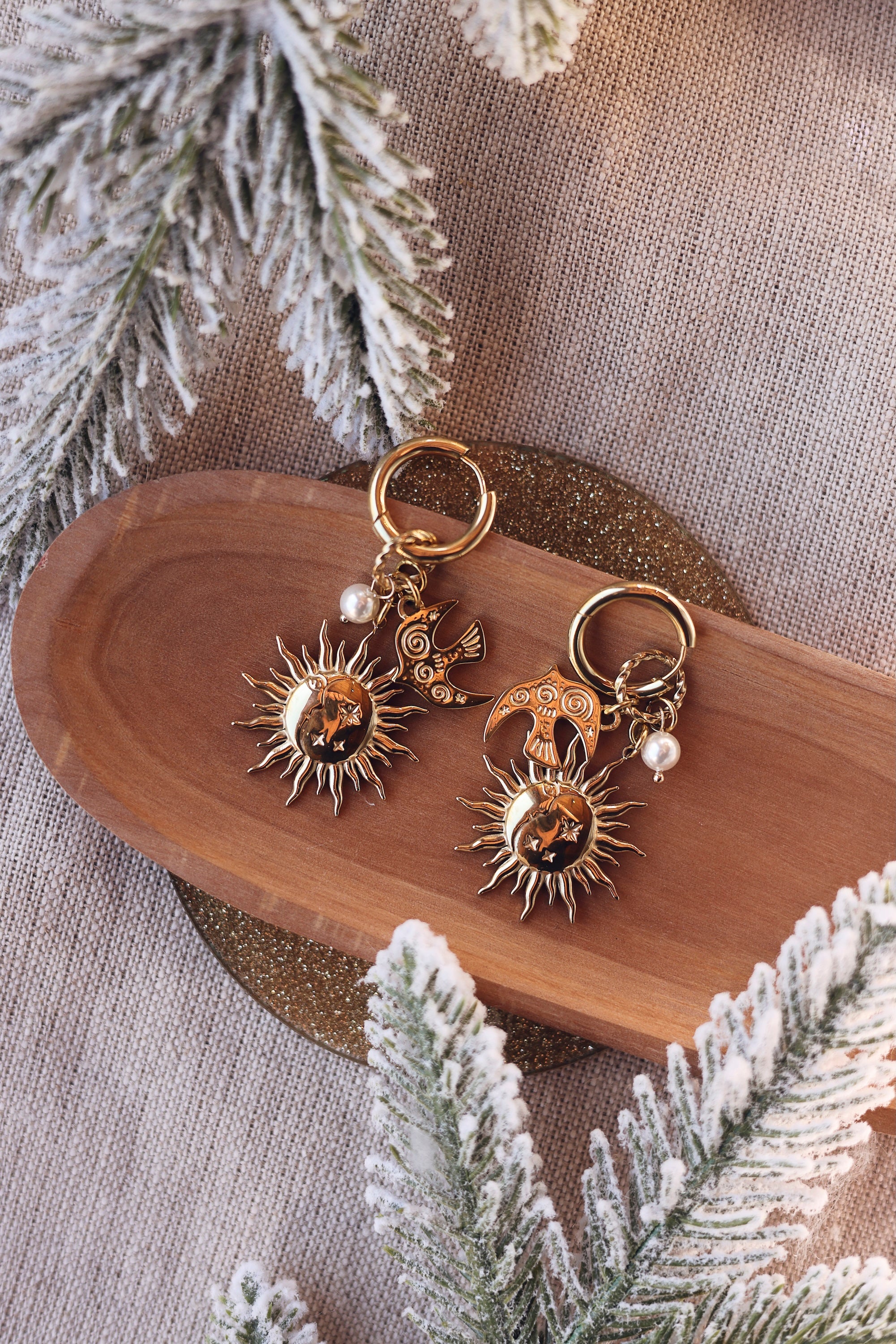 Boucles d'oreilles mini créoles dorées Solenia pendentif soleil, lune, nacre et hirondelle - Bijou photographié sur une coupole en bois avec des branches de sapin