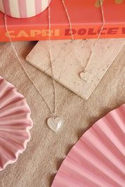 Médaille en nacre Gina M avec bordure en argent en forme de cœur pour gravure - Bijou photographié sur du tissu en lin beige avec des décorations oranges et roses