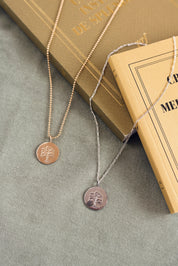 Médaille réversible dorée Arbre de vie avec une face ornée d'un arbre de vie et une face lisse pour gravure - Bijou photographié sur un tissu kaki avec des livres