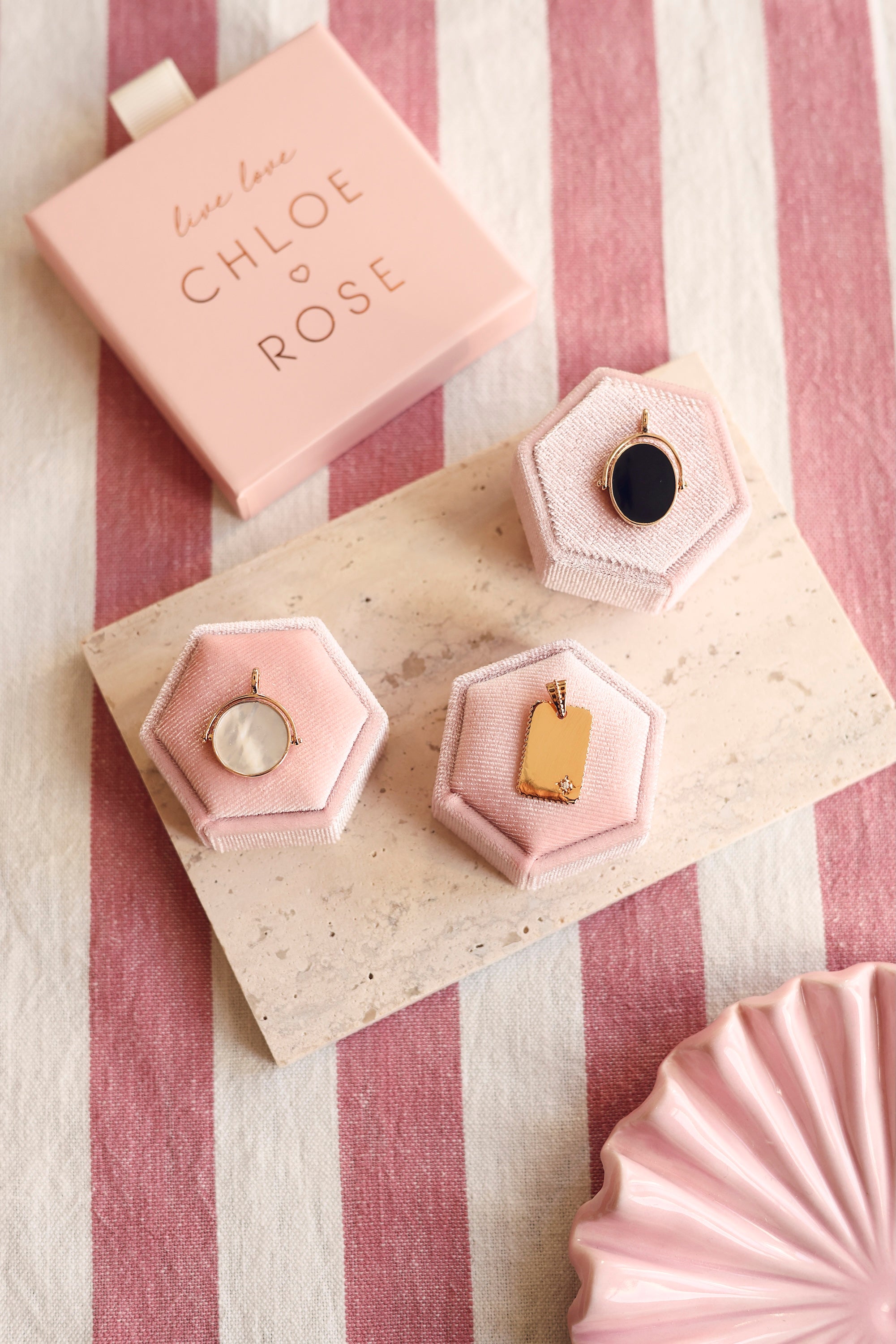 Médaille réversible ronde dorée Joy avec un côté en nacre et un côté à graver - Bijou photographié sur un présentoir rose sur une pierre rectangulaire avec des décorations roses et une boîte chloe rose