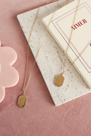 Médaille rectangulaire Sarah dorée avec bordure sertie de zircons pour gravure - Bijou photographié sur un tissu rose avec une planche en marbre et un livre