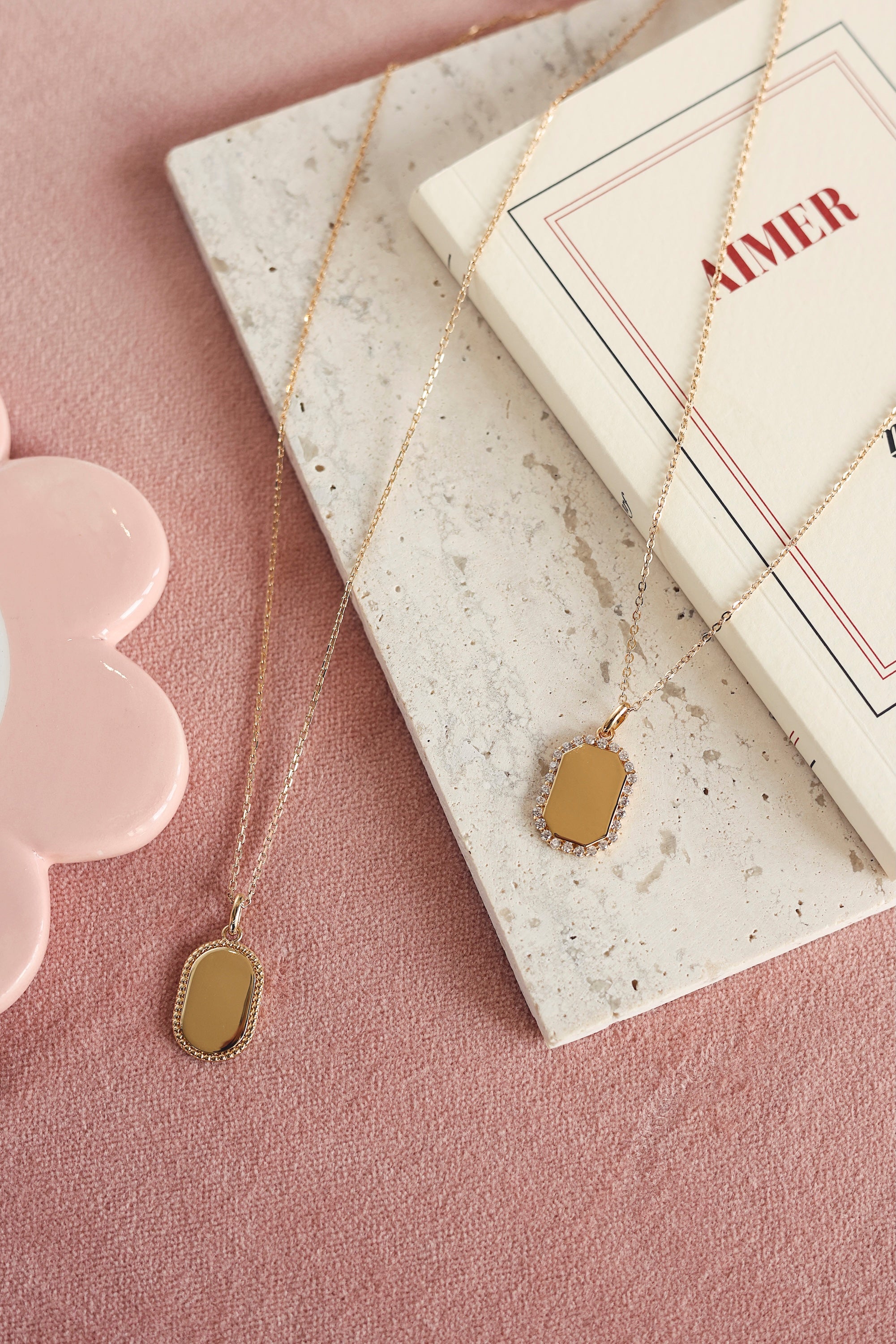 Médaille rectangulaire Sarah dorée avec bordure sertie de zircons pour gravure - Bijou photographié sur un tissu rose avec une planche en marbre et un livre